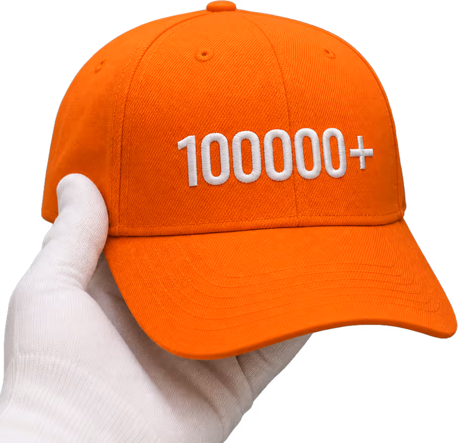 Orange baseball cap with the word 'KAPTURE' embroidered on the front in white, held by an industrial embroidery machine.