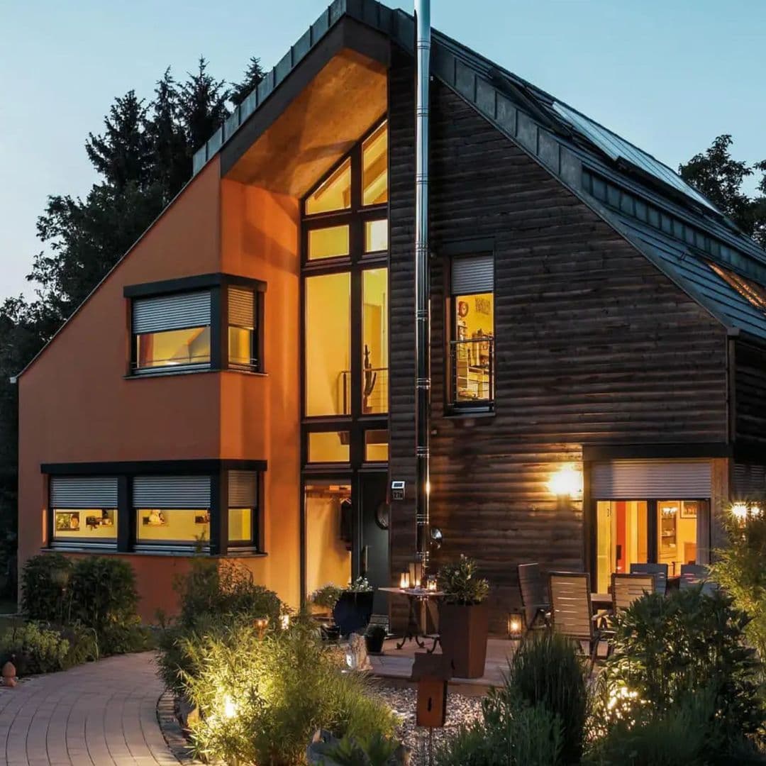 Modernes Haus mit orangefarbener und Holz-Fassade, beleuchtet am Abend mit Garten und Außenmöbeln.