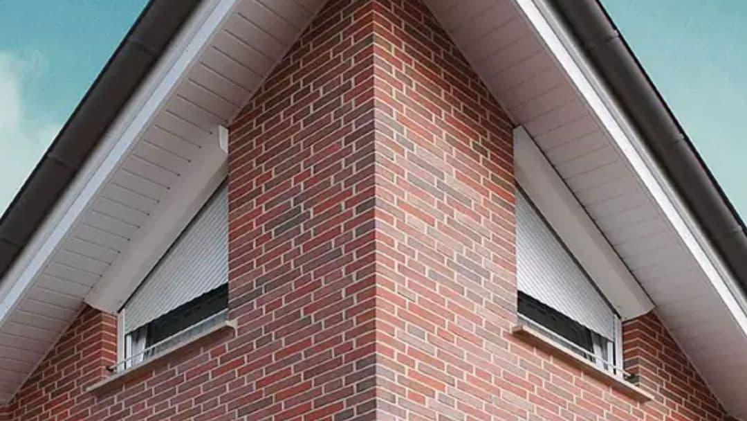 Eckansicht eines Backsteinhauses mit zwei Fenstern, die geschlossene weiße Rollläden haben.