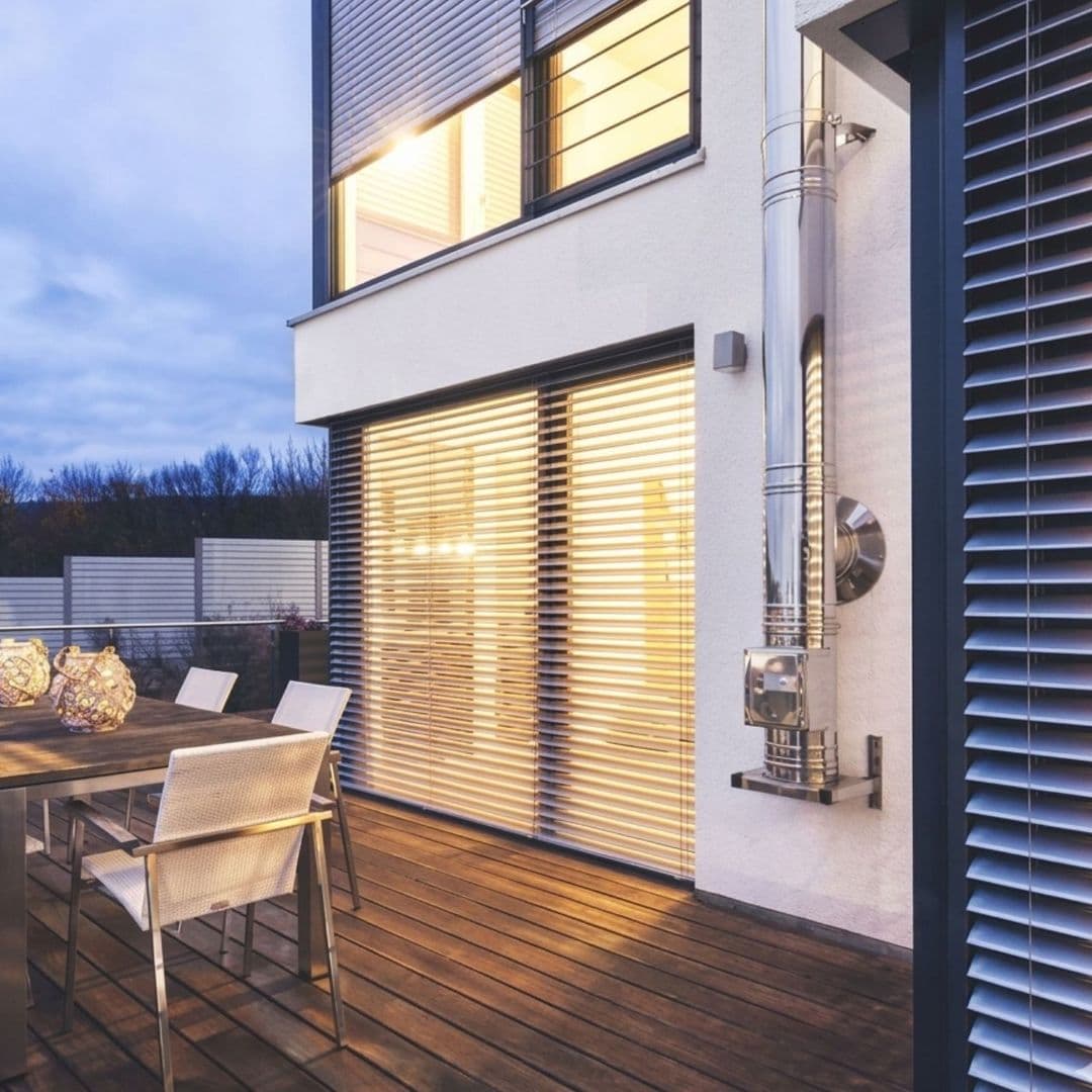 Moderne Terrasse mit Holzboden, Gartentisch und Stühlen neben einem Haus mit geschlossenen Außenjalousien bei Dämmerung.
