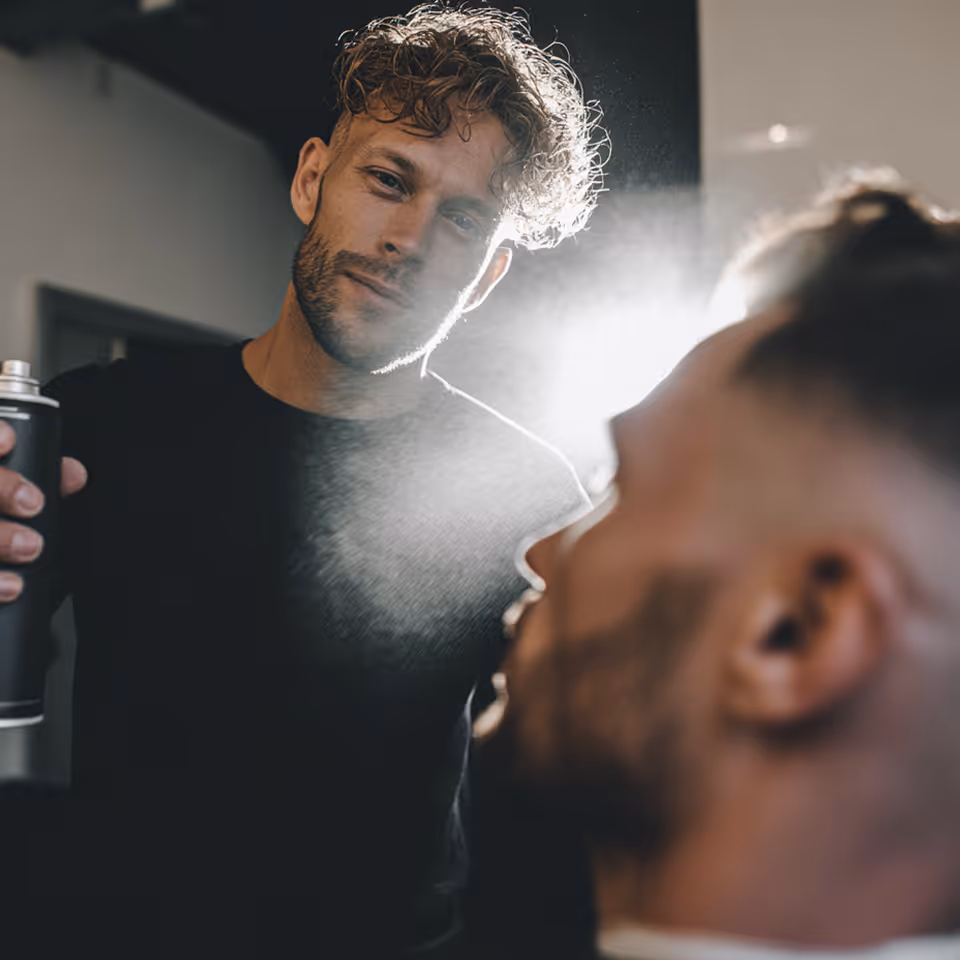 Peluquero con barba rociando spray sobre el cabello de un cliente con corte de barba en un salón.
