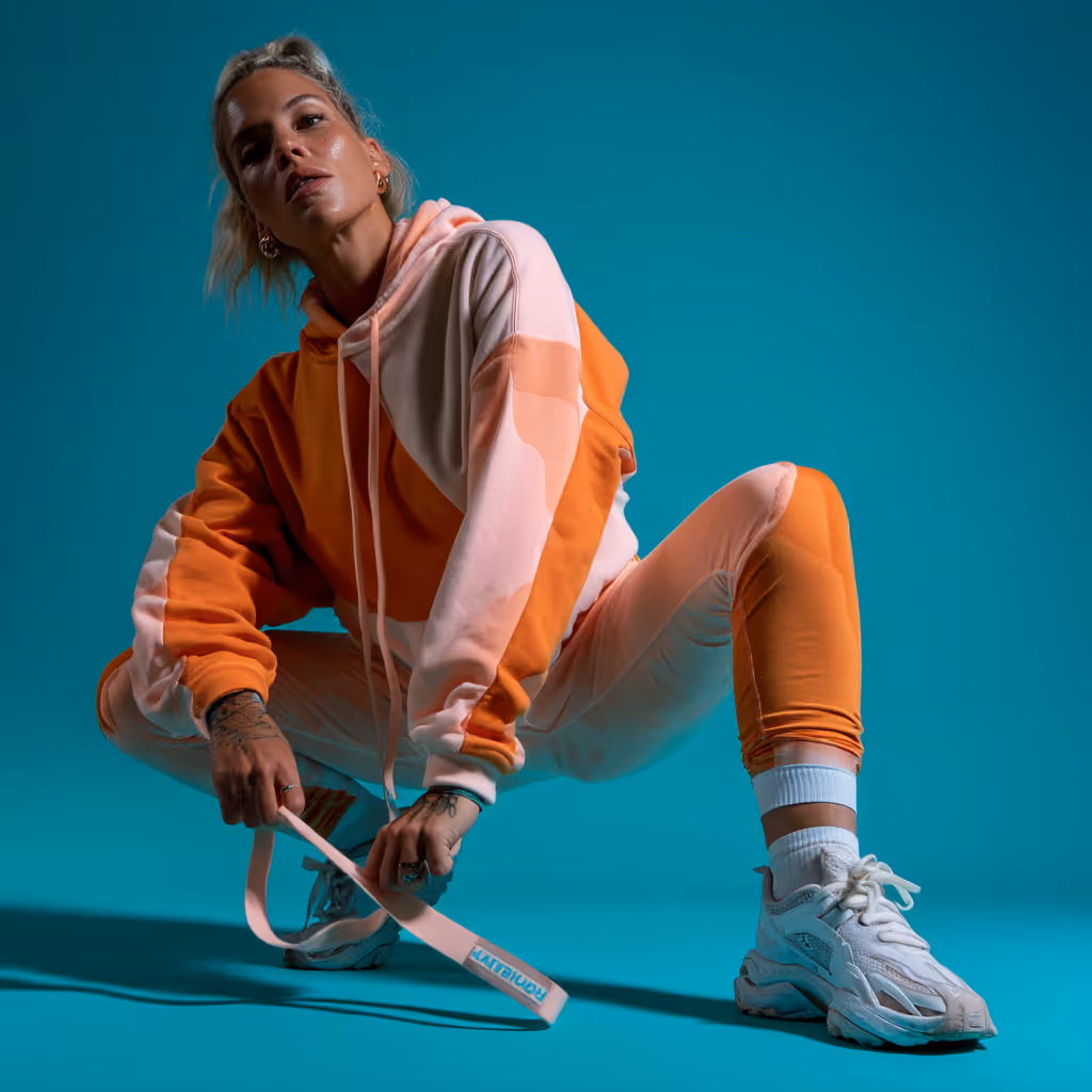 Mujer rubia con tatuajes usando chándal naranja y rosa, agachada en cuclillas con fondo azul.