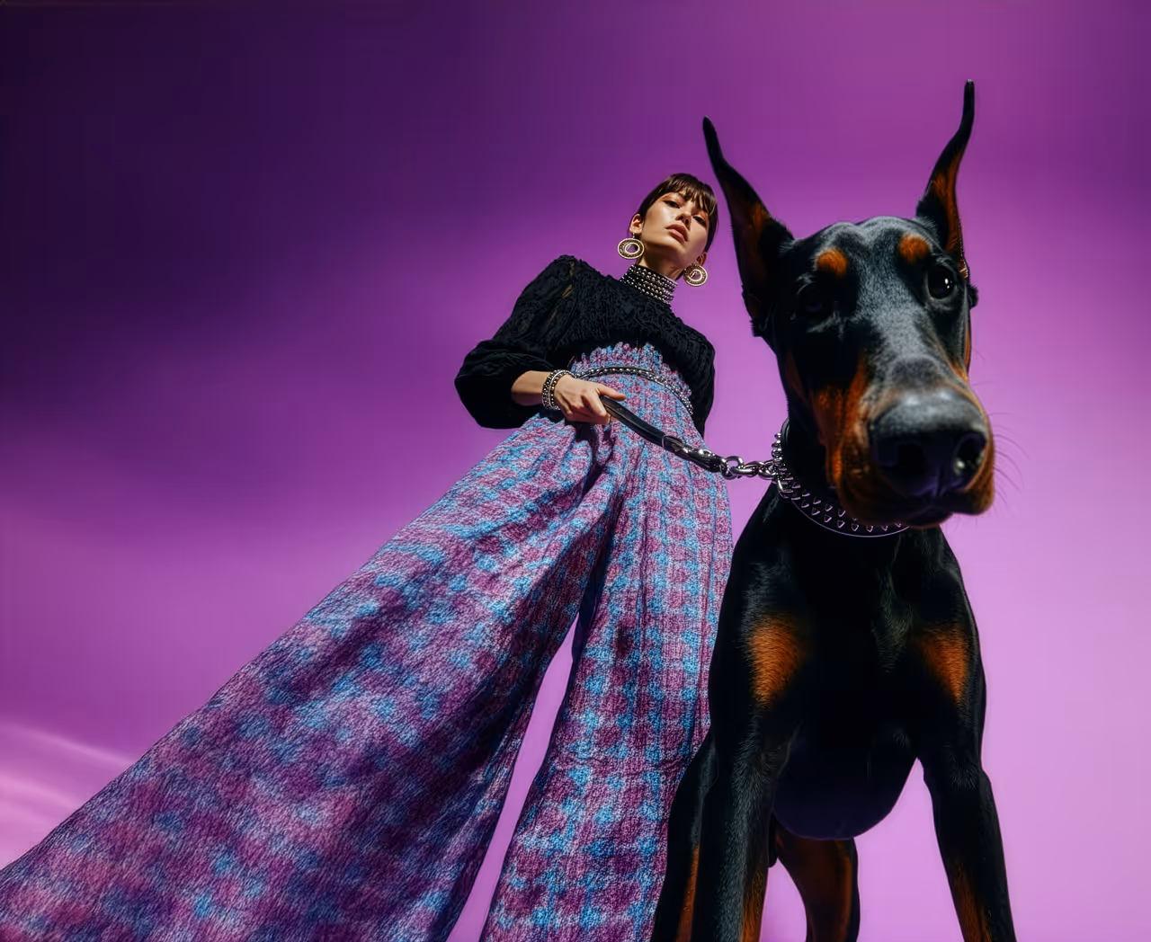 Low-angle view of a woman in a purple patterned wide-leg pants and black top holding the leash of a large Doberman dog against a purple background.