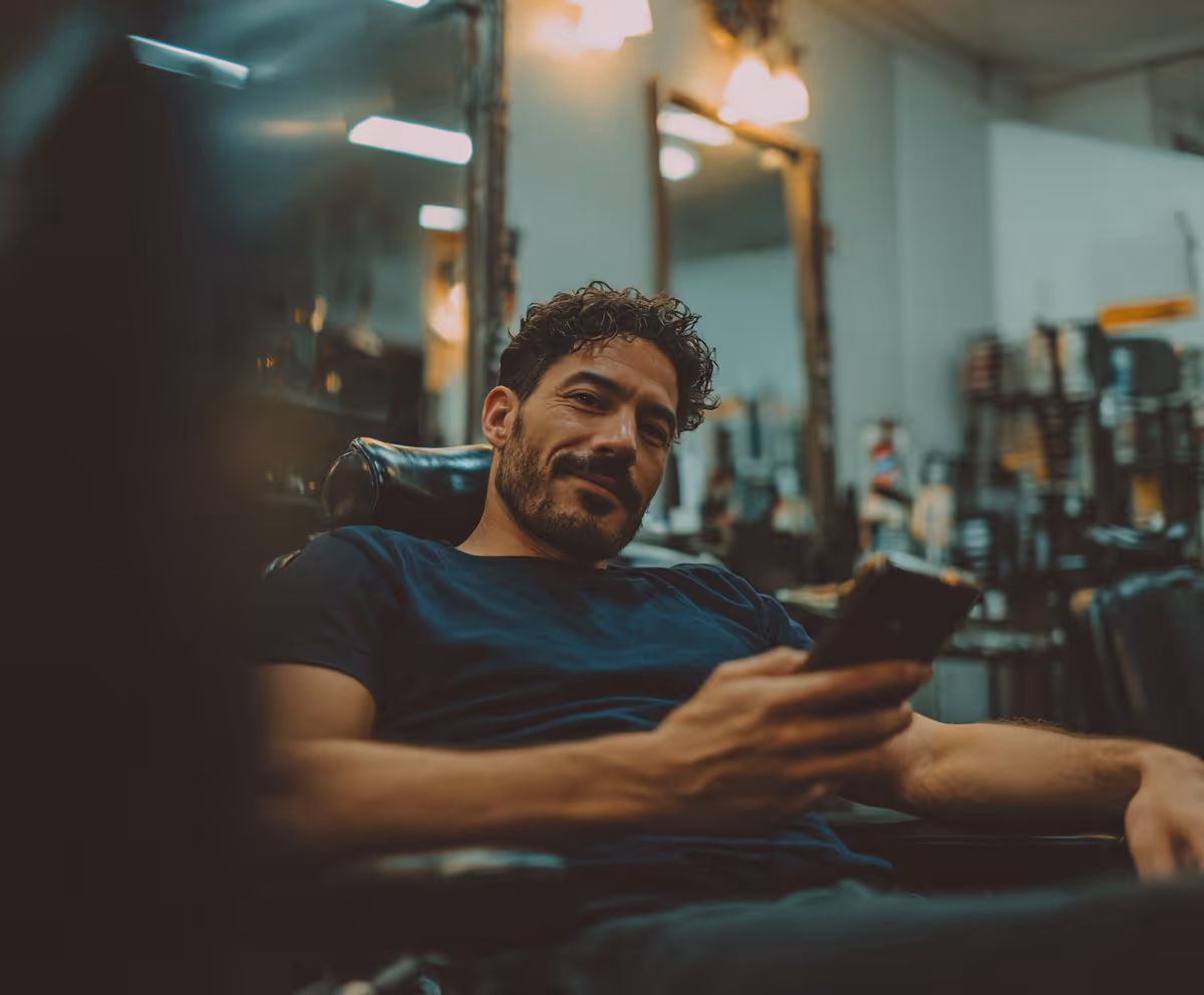 Man with curly hair and beard sitting relaxed in a barber chair, holding a smartphone in a barbershop.