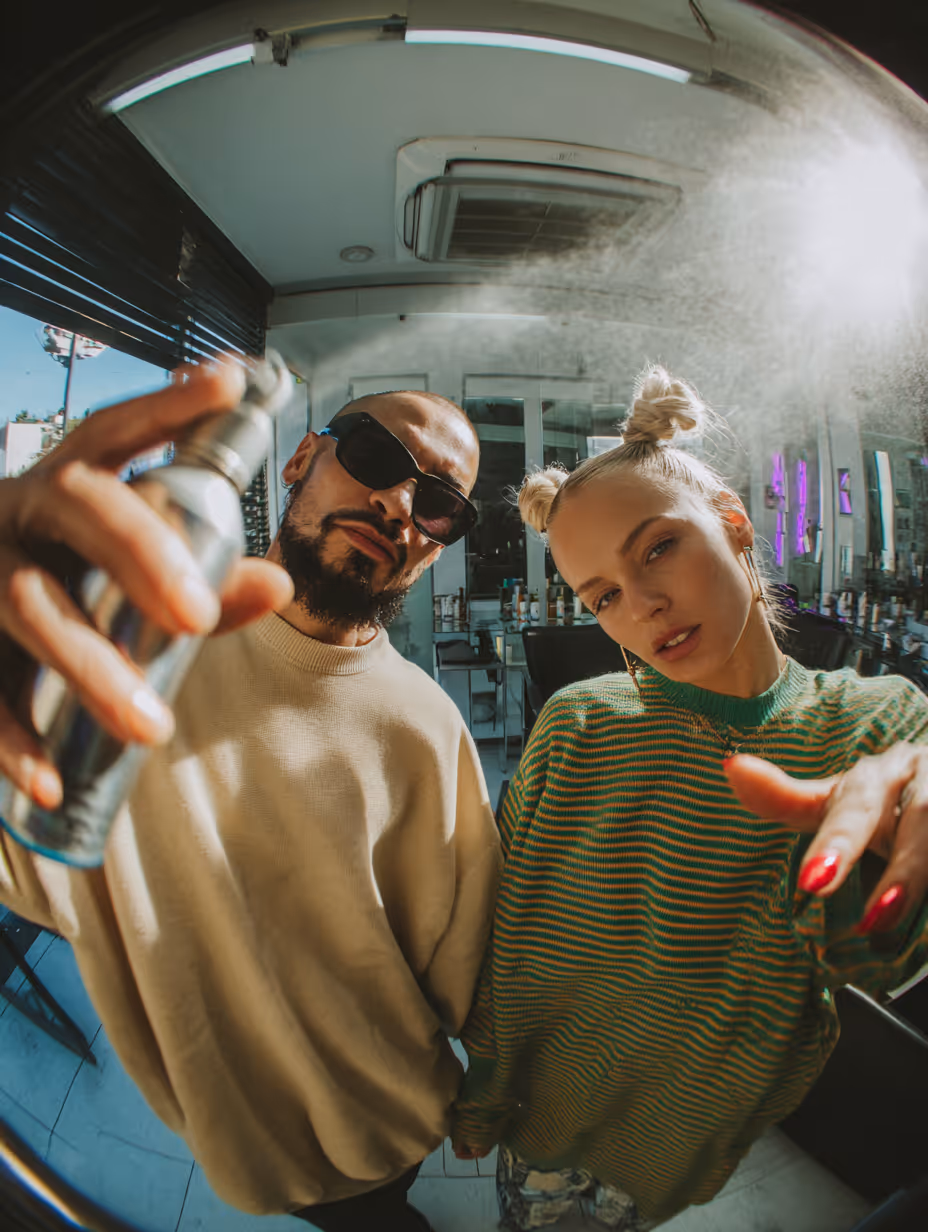 Man wearing sunglasses sprays hair spray while woman with blonde space buns and green striped sweater poses in a salon.