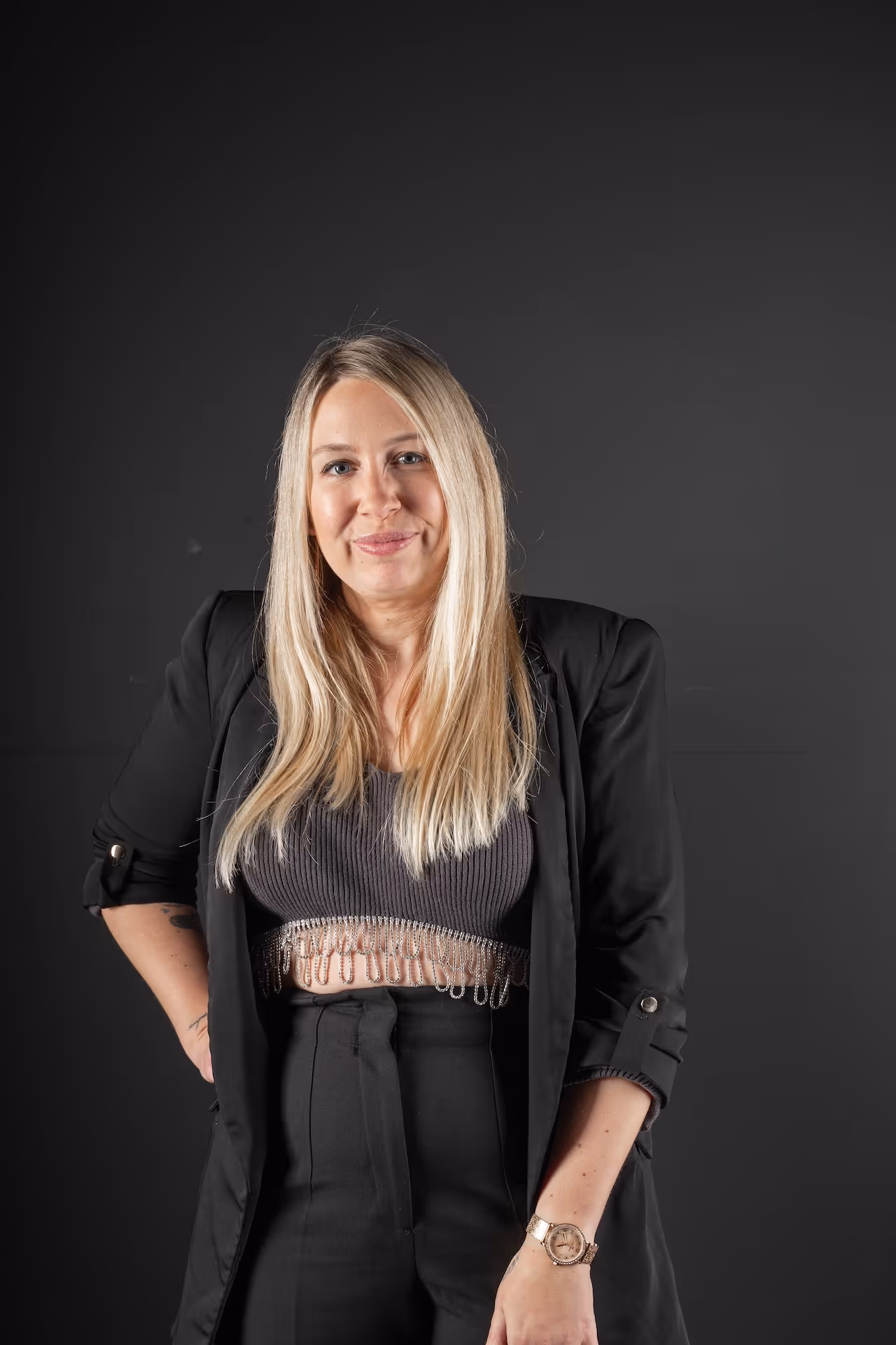 Mujer rubia sonriendo con chaqueta negra con lentejuelas y pantalones negros frente a fondo gris claro.