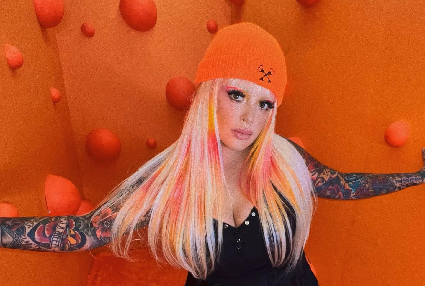 Person with long blonde hair streaked with pink and orange, wearing an orange beanie and black top, with tattooed arms against an orange background with bubble-like shapes.