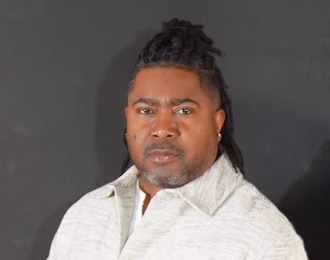Man with medium-length dreadlocks wearing a white collared shirt against a dark background.