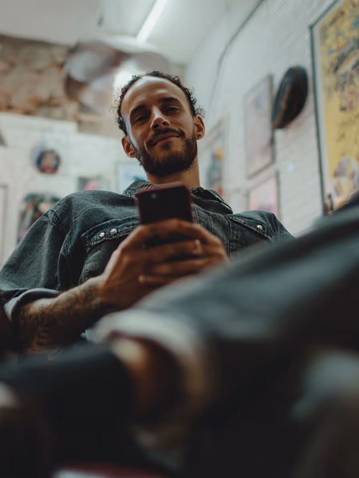 Bearded man in denim shirt holding a smartphone, viewed from a low angle indoors.