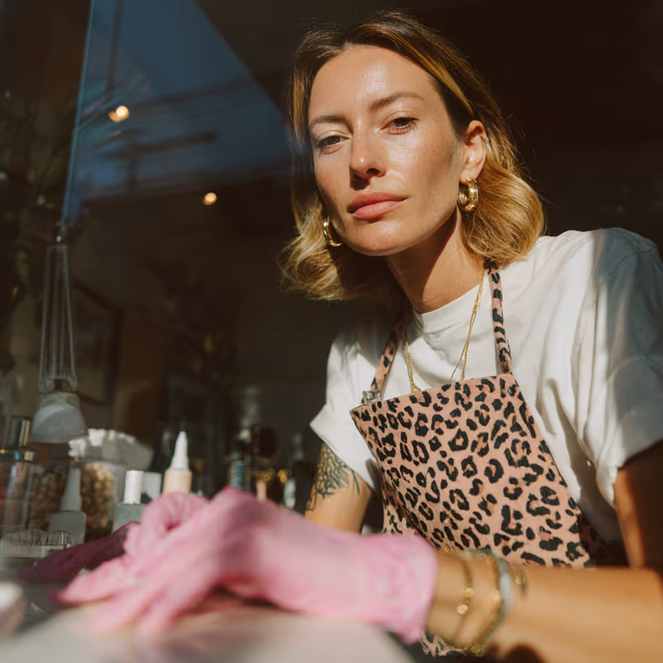 Woman wearing pink gloves and leopard print apron, leaning forward with a focused expression in a well-lit room.