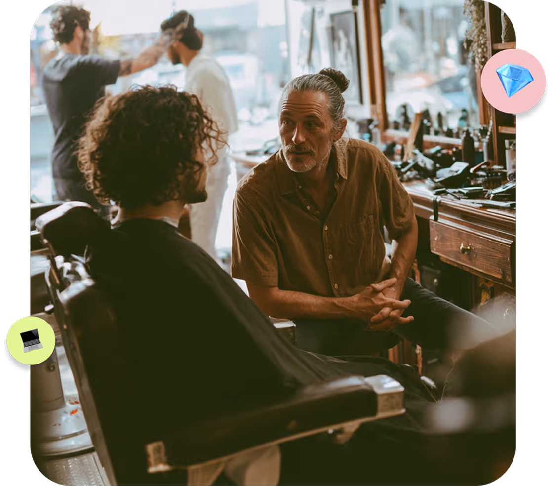 Barber in a brown shirt talking to a seated customer with curly hair in a barbershop.