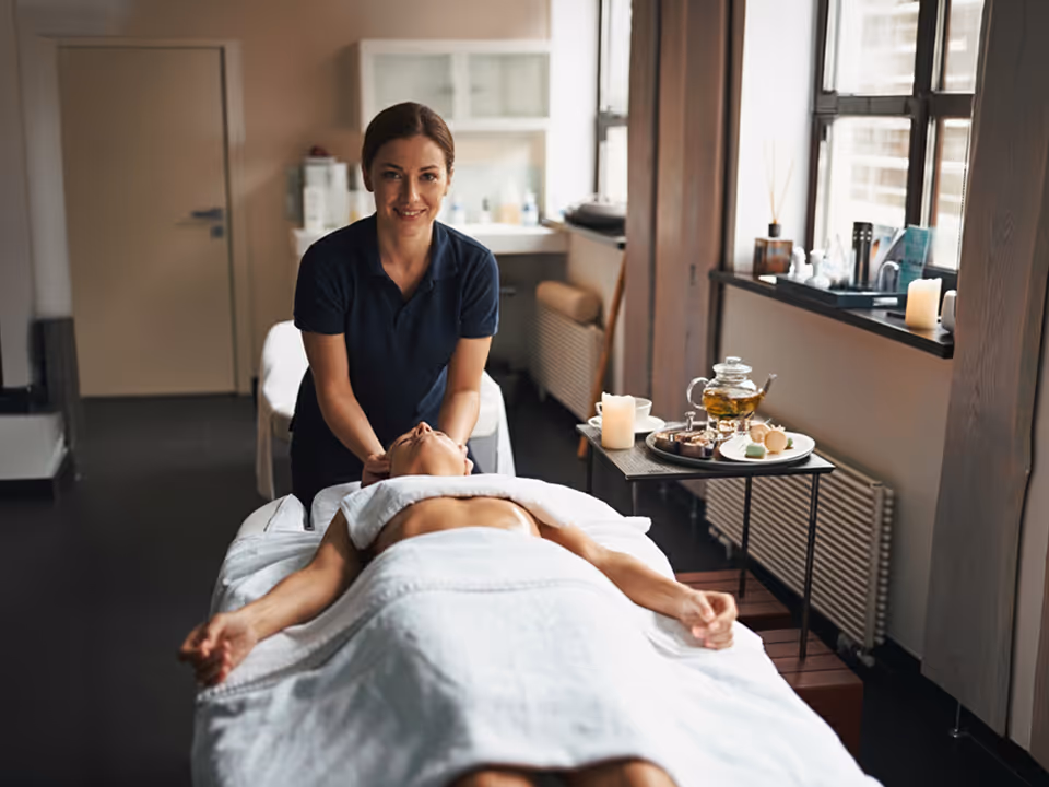 Masażystka w ciemnym ubraniu masuje szyję kobiety leżącej na stole do masażu w jasnym, przytulnym gabinecie spa.