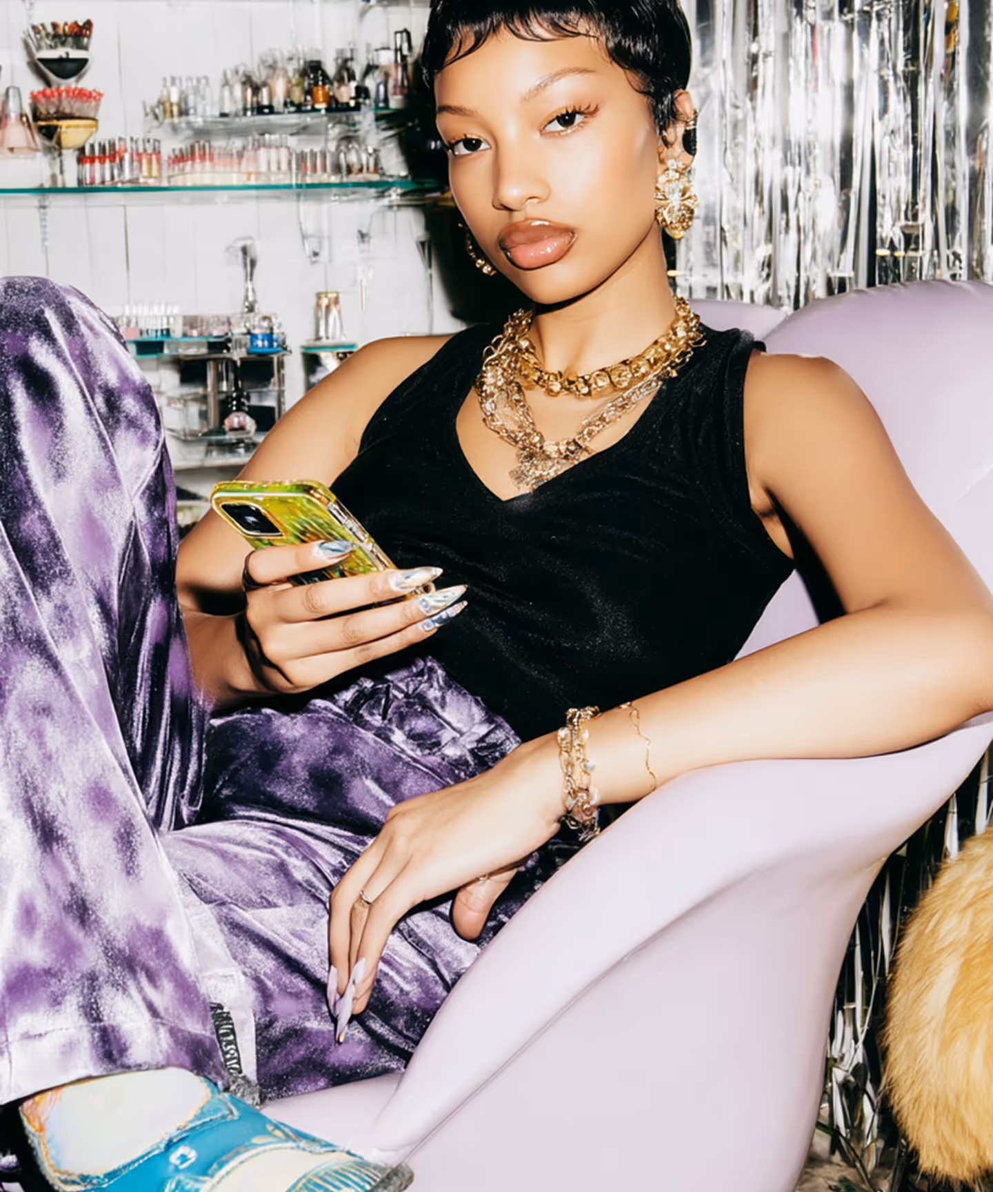 Młoda kobieta z krótkimi włosami, w czarnej koszulce i fioletowych spodniach, siedzi na jasnoróżowym fotelu i trzyma telefon.