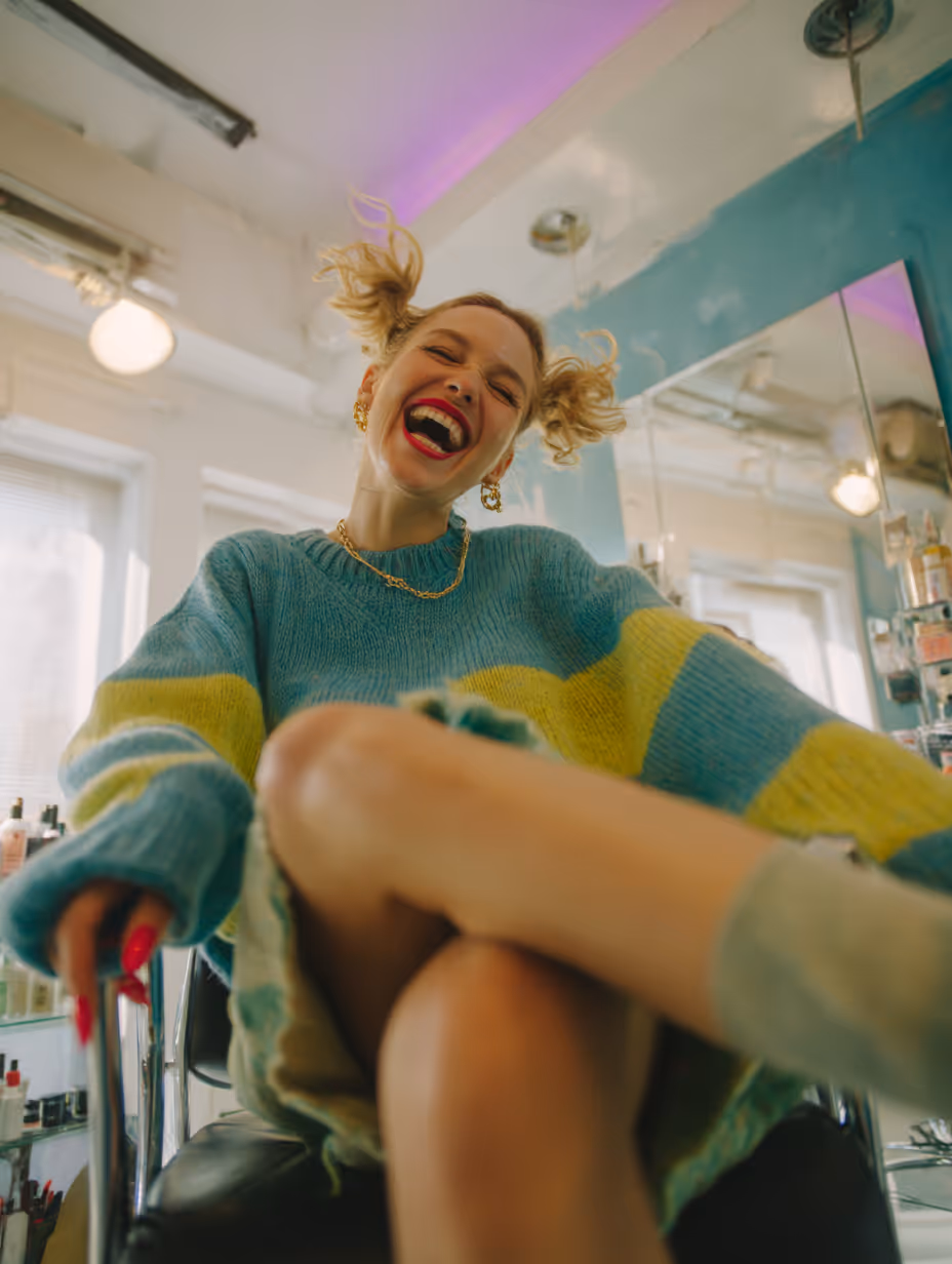 Młoda kobieta z lekkimi loczkami i jasnoczerwonymi ustami śmiejąca się radośnie, siedząc na krześle w kolorowym swetrze w jasnym pokoju.