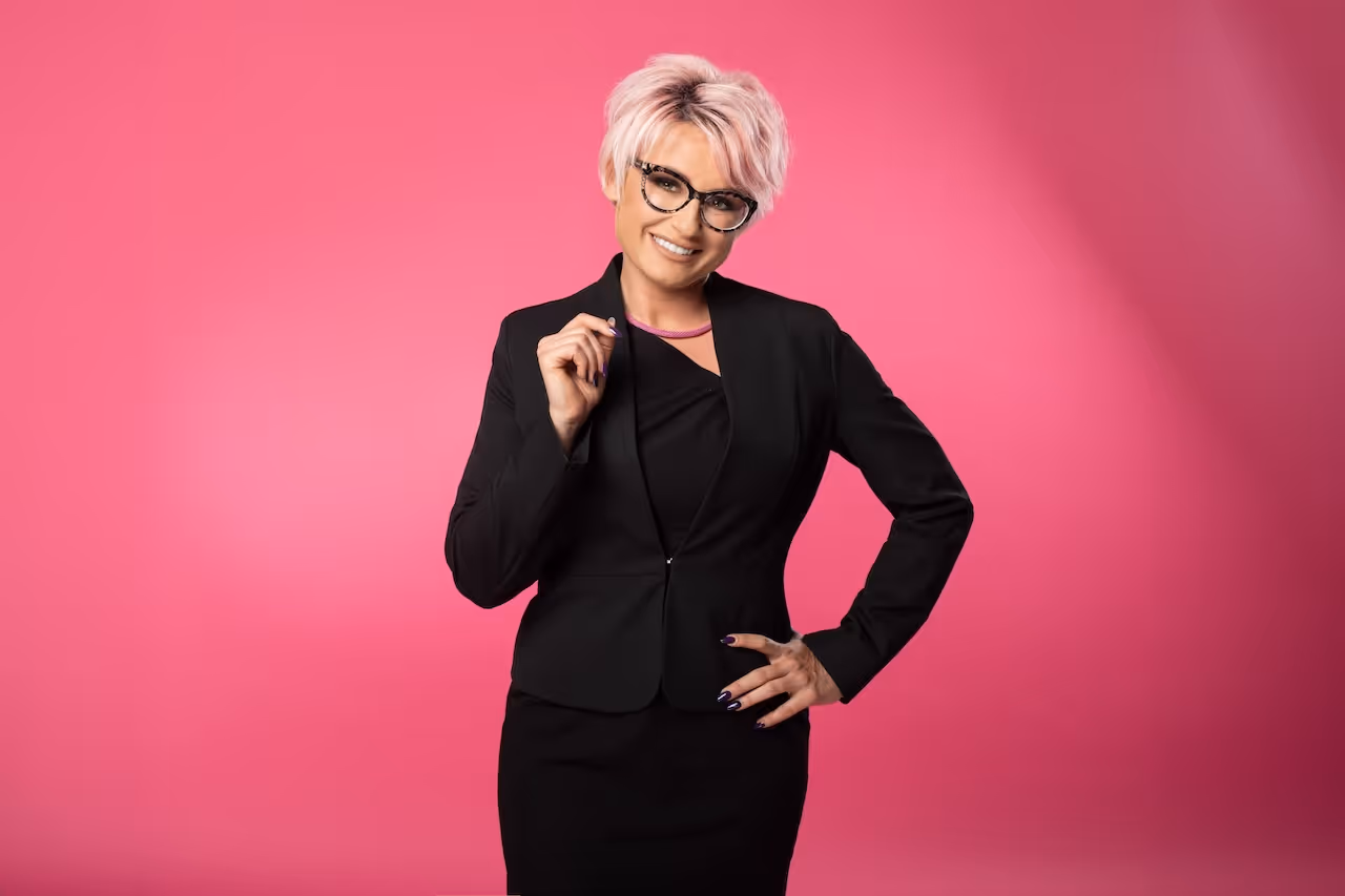Kobieta z krótkimi różowymi włosami, uśmiechająca się, ubrana w czarną marynarkę z głębokim dekoltem i delikatnym naszyjnikiem.