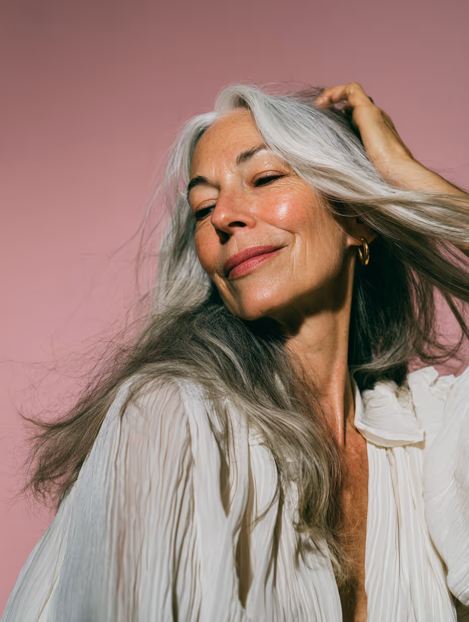 Uśmiechnięta starsza kobieta z długimi siwymi włosami, ubrana w białą plisowaną bluzkę na różowym tle.
