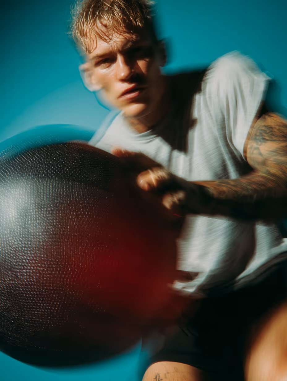 Mężczyzna z tatuażami ćwiczący z dużą czarną piłką fitness na turkusowym tle.