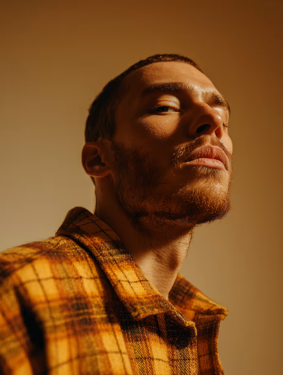 Mężczyzna z krótkimi włosami i zarostem w pomarańczowej koszuli w kratę patrzący pewnie w kamerę pod kątem.