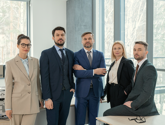 Five professionals in business attire standing and sitting in a modern office with large windows.