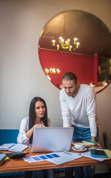 Two coworkers reviewing something on a laptop at a table with papers, books, and coffee cups in a modern office.