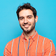 Smiling man with dark hair and a beard wearing an orange striped shirt against a blue background.