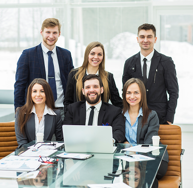 Five smiling business professionals gathered around a laptop at a glass conference table with documents.