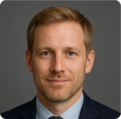 Headshot of a smiling Caucasian man with short light brown hair and blue eyes wearing a suit and tie against a gray background.