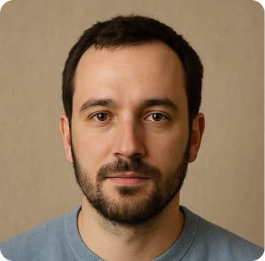 Close-up portrait of a man with short dark hair, a beard, and brown eyes wearing a blue sweater against a beige background.