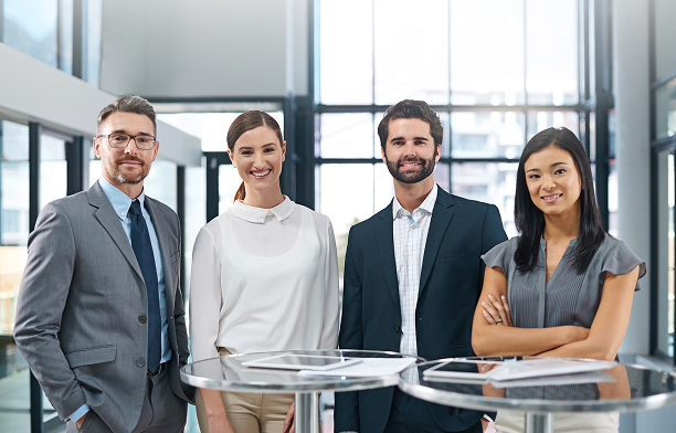 Four professionally dressed colleagues standing together behind a table in a modern office.