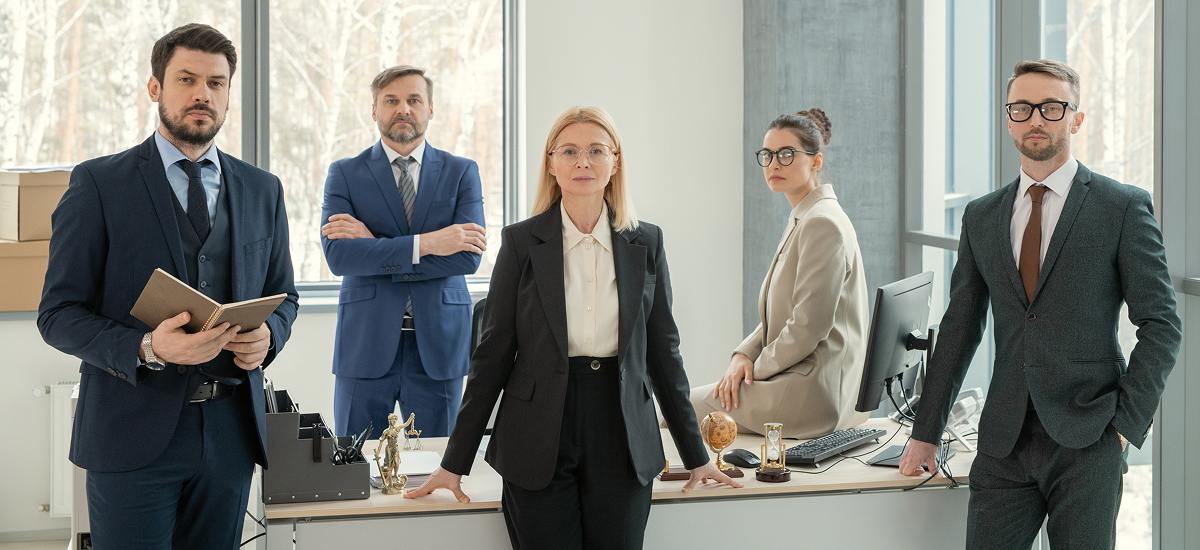 Five professional businesspeople in suits posing confidently in a modern office with large windows and desks.