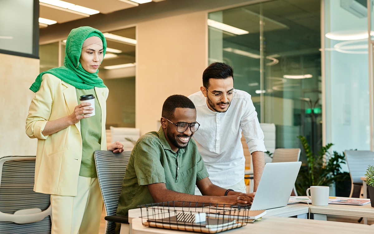 Three colleagues in an office collaborating around a laptop, one seated and two standing, with one holding a coffee cup.