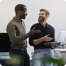 Two men having a discussion, one holding a laptop, in a modern office setting.