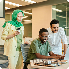 Three diverse coworkers collaborating and smiling in a modern office, one seated at a laptop and two standing nearby.