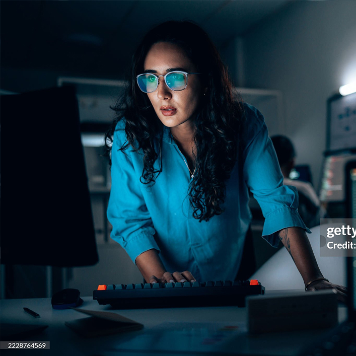 Frau mit Brille und blauem Hemd, die konzentriert vor einem Computerbildschirm in einem dunklen Büro arbeitet.