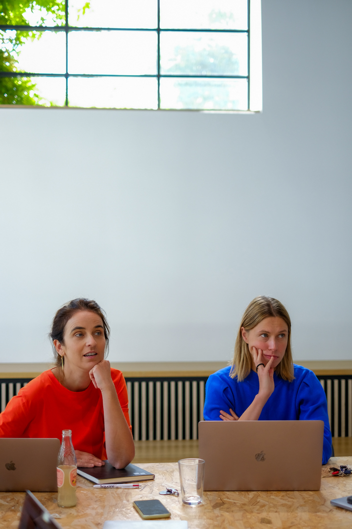 Twee vouwen zitten naast elkaar aan de bureau. Een heeft een rode shirt, de andere een blauwe. Het is teamoverleg.