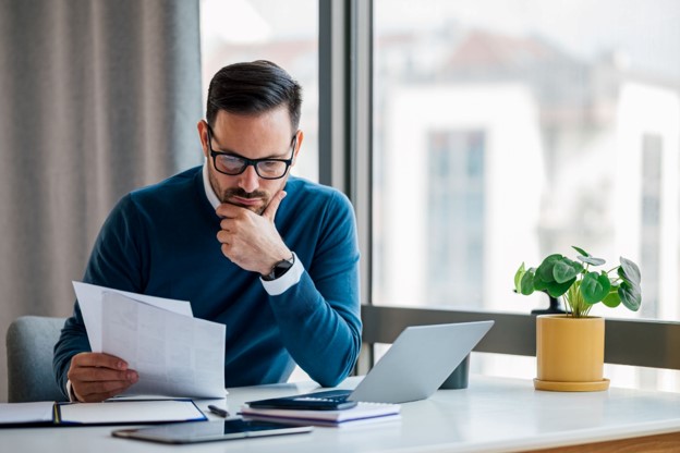 Man reviewing freight invoice documents beside a laptop while checking paperwork for billing accuracy.