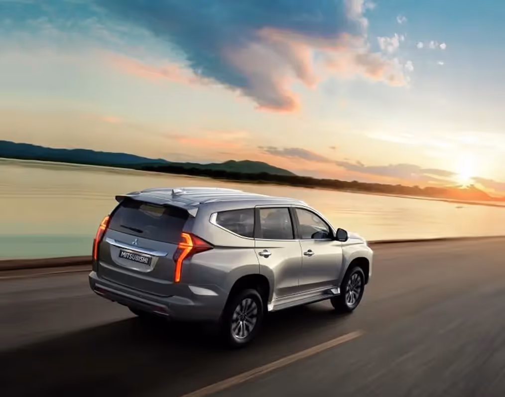 Silver Mitsubishi SUV driving on a road beside a large body of water at sunset with mountains in the background.
