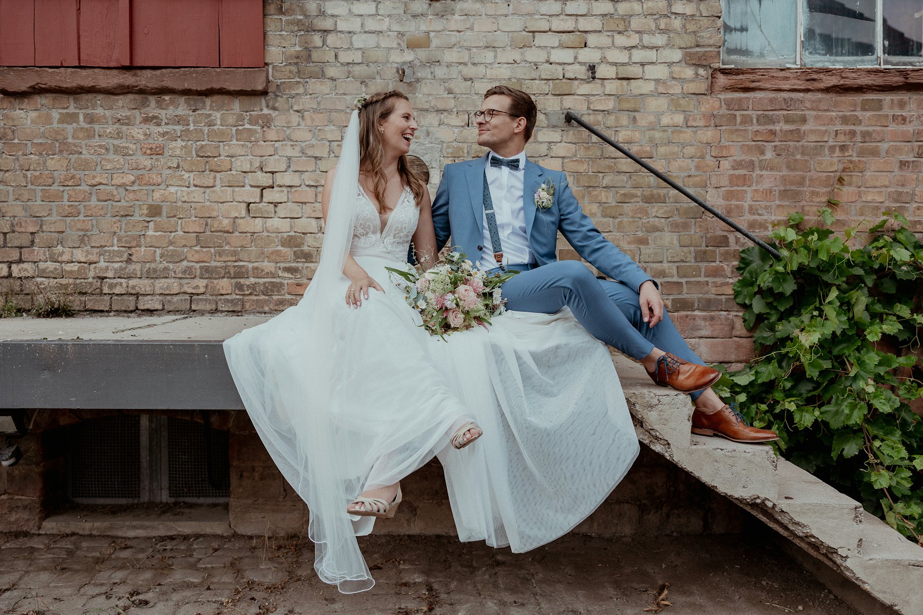 Braut und Bräutigam im blauen Anzug sitzen nach ihrer Hochzeit in Frankfurt auf einer Backsteintreppe