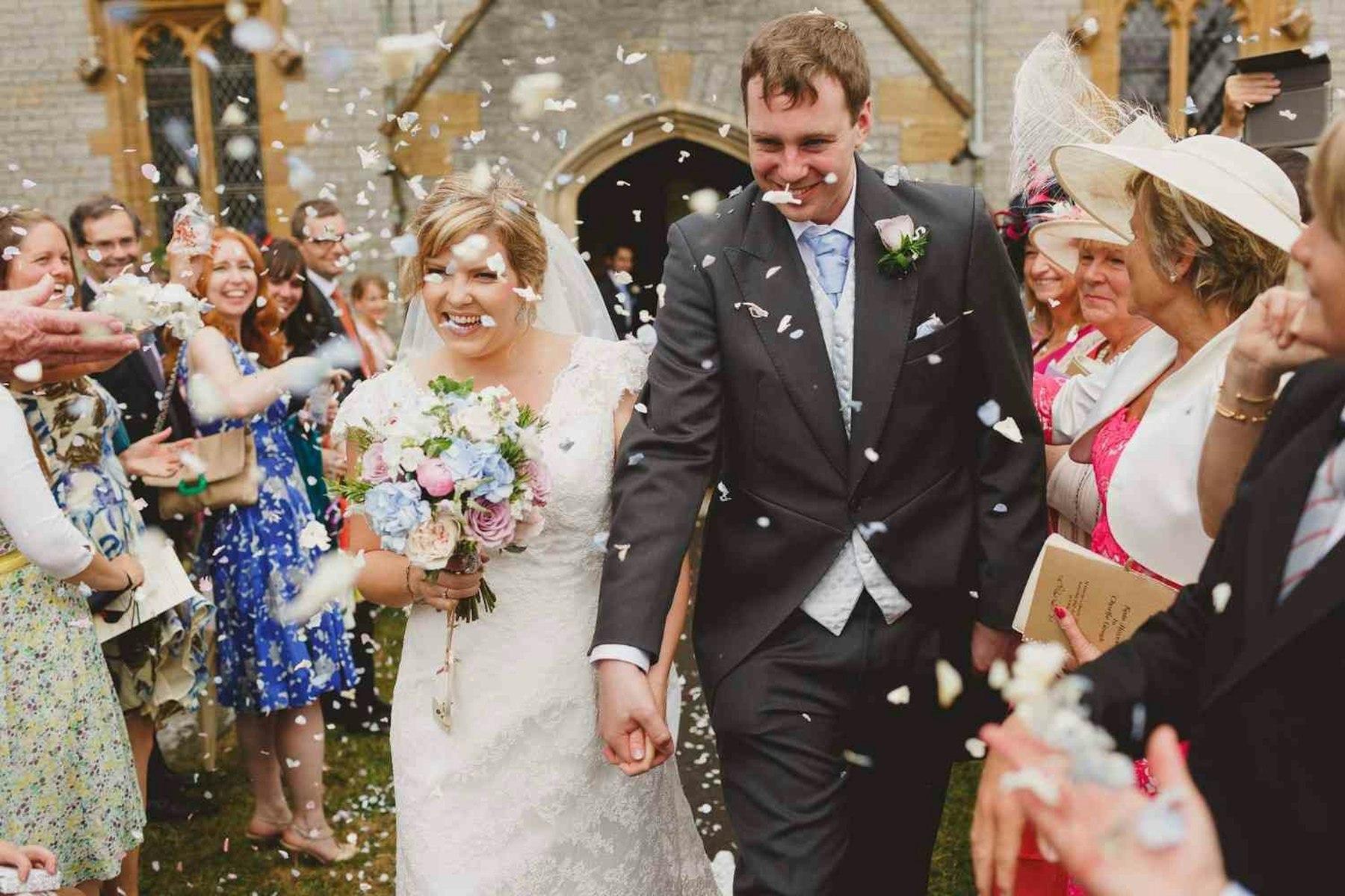 Guests in hats and morning suits throw confetti over a bride and groom leaving a village church in Somerset