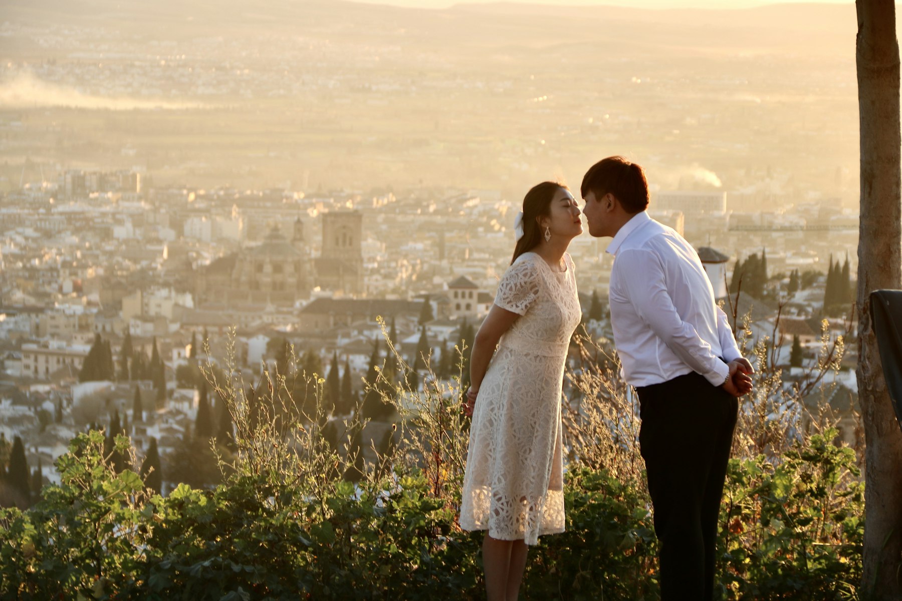 Una pareja con vestido de encaje blanco y camisa blanca al atardecer en un mirador sobre Granada