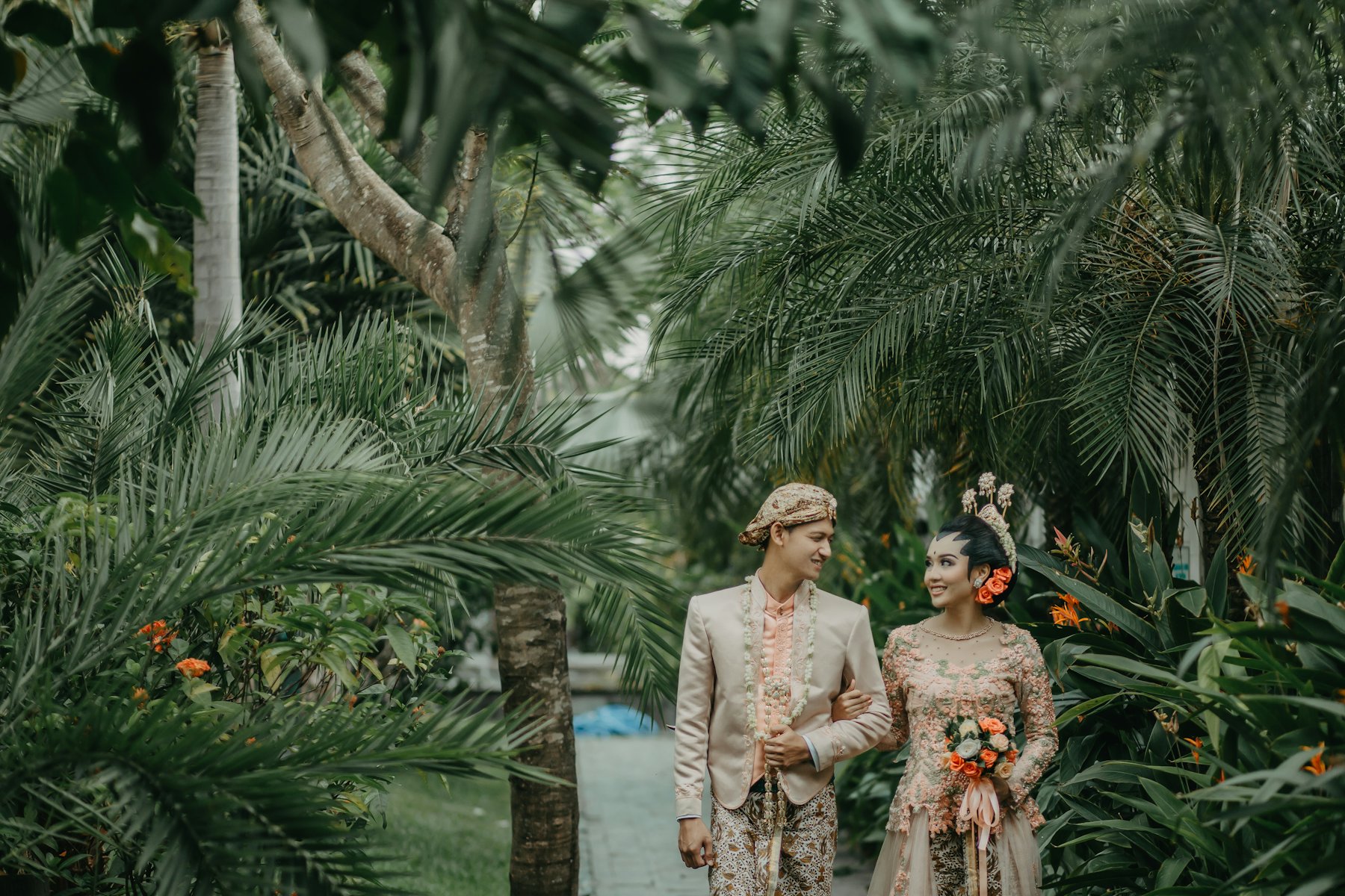 An Indonesian bride and groom in traditional Javanese kebaya, batik and jasmine garlands walk arm in arm through a tropical garden