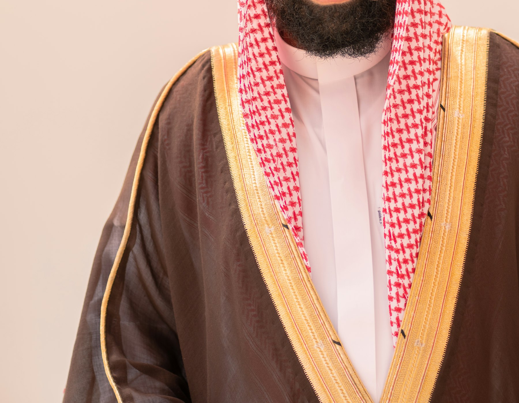 Ein Mann in einem goldbesetzten Bisht über einem weißen Thobe mit rotem Shemagh, festliche Kleidung bei saudischen Hochzeiten