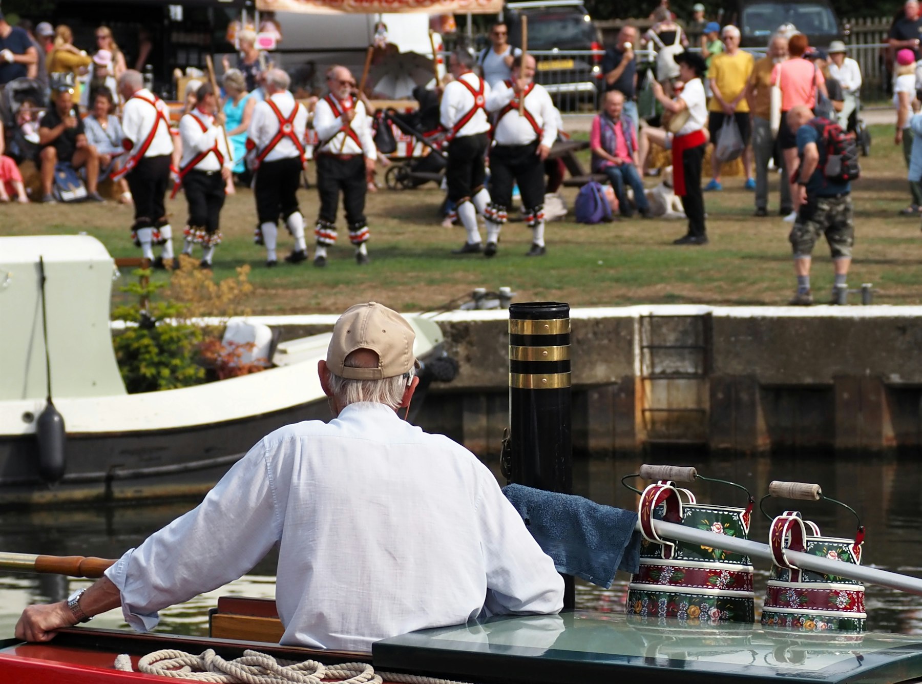 Morris-Tänzer in weißen Hemden mit roten Schärpen und Schellen tanzen auf einer Wiese am Fluss in Guildford, gesehen von einem Narrowboat mit bemaltem Kanalgeschirr
