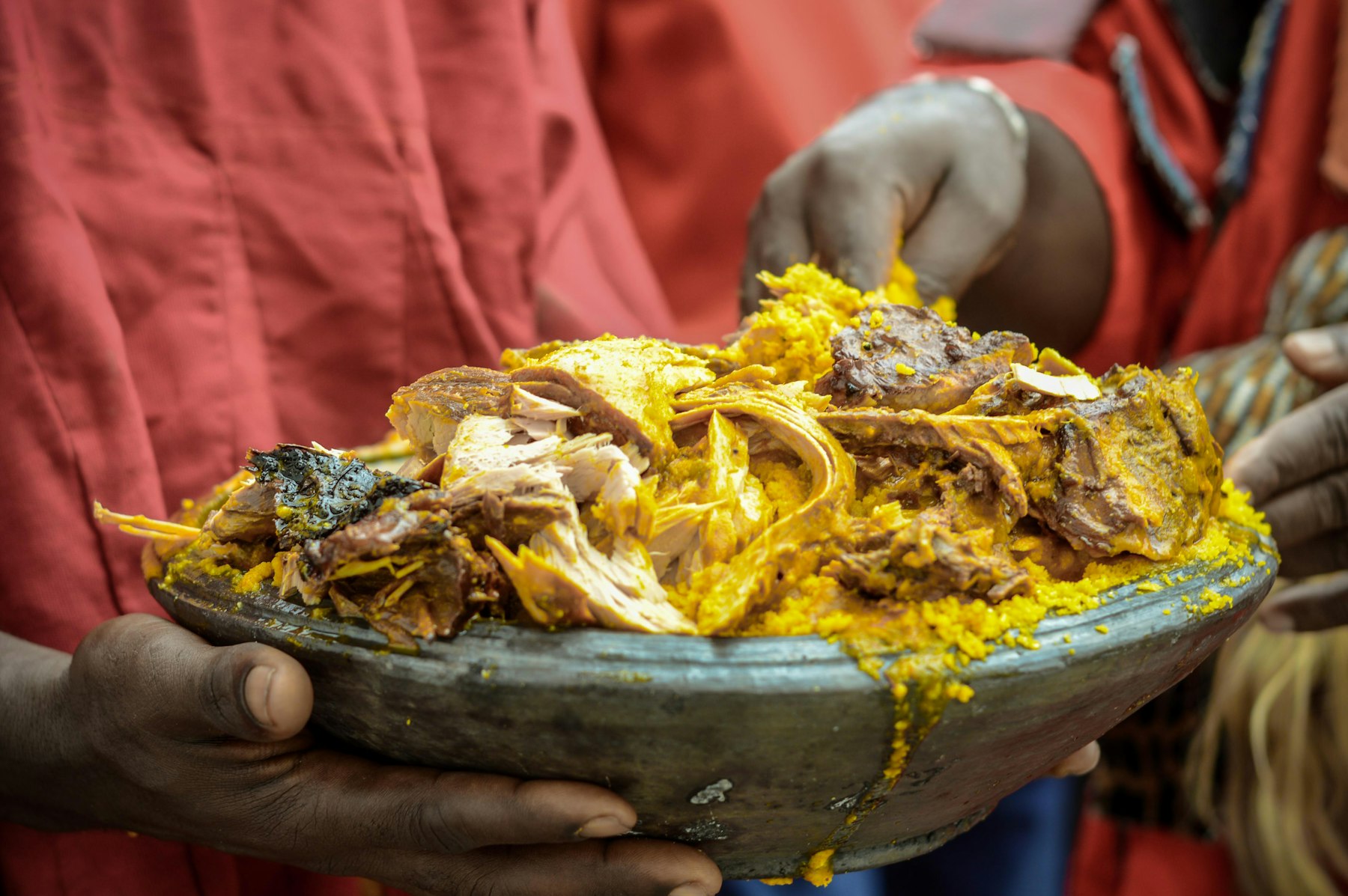 Hands holding a clay bowl of kpokpoi, the yellow cornmeal dish of Homowo, with palm-nut soup and fish