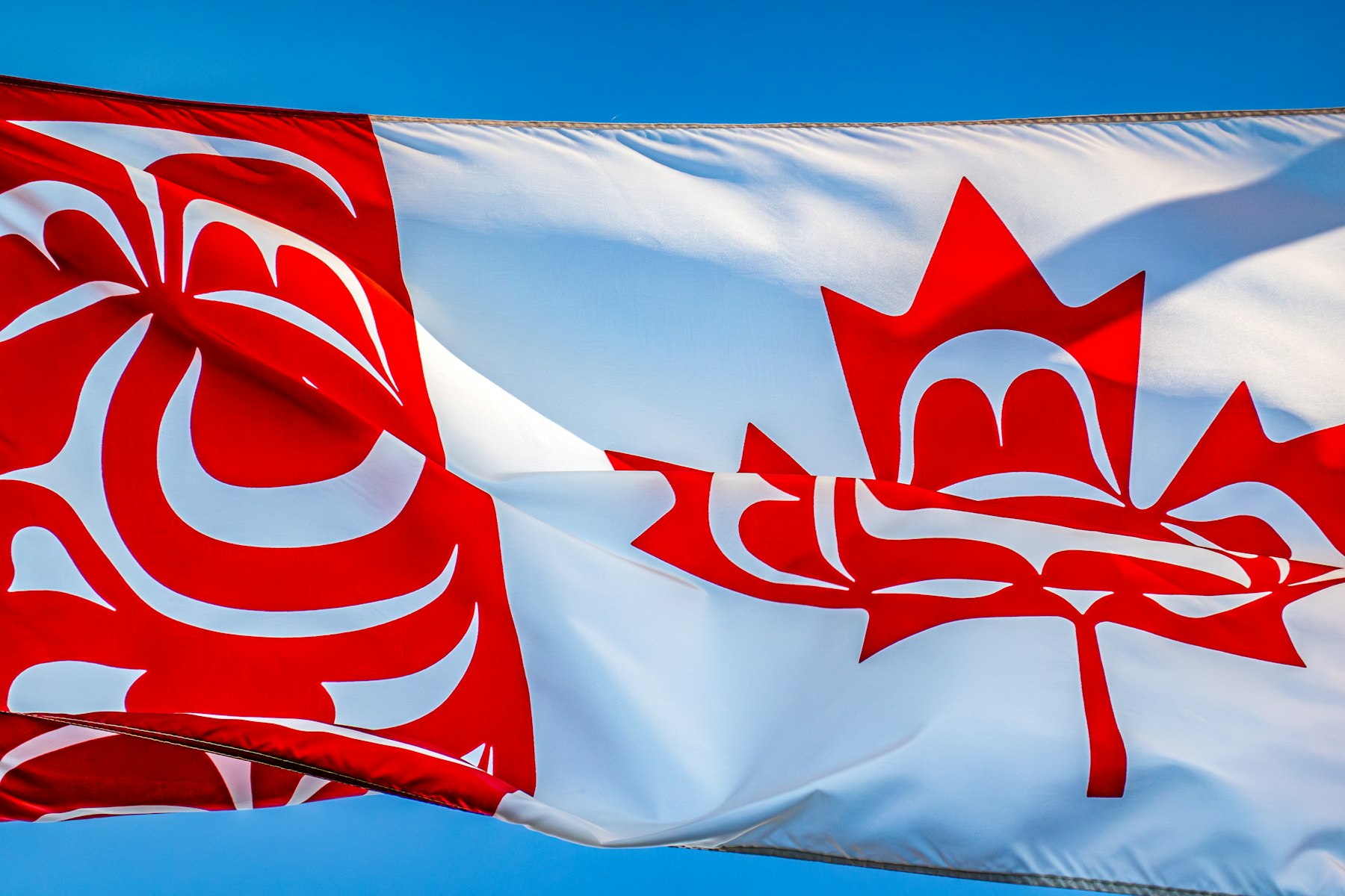 Una bandera de Canadá reinterpretada con arte formline de las Primeras Naciones, ondeando bajo un cielo azul