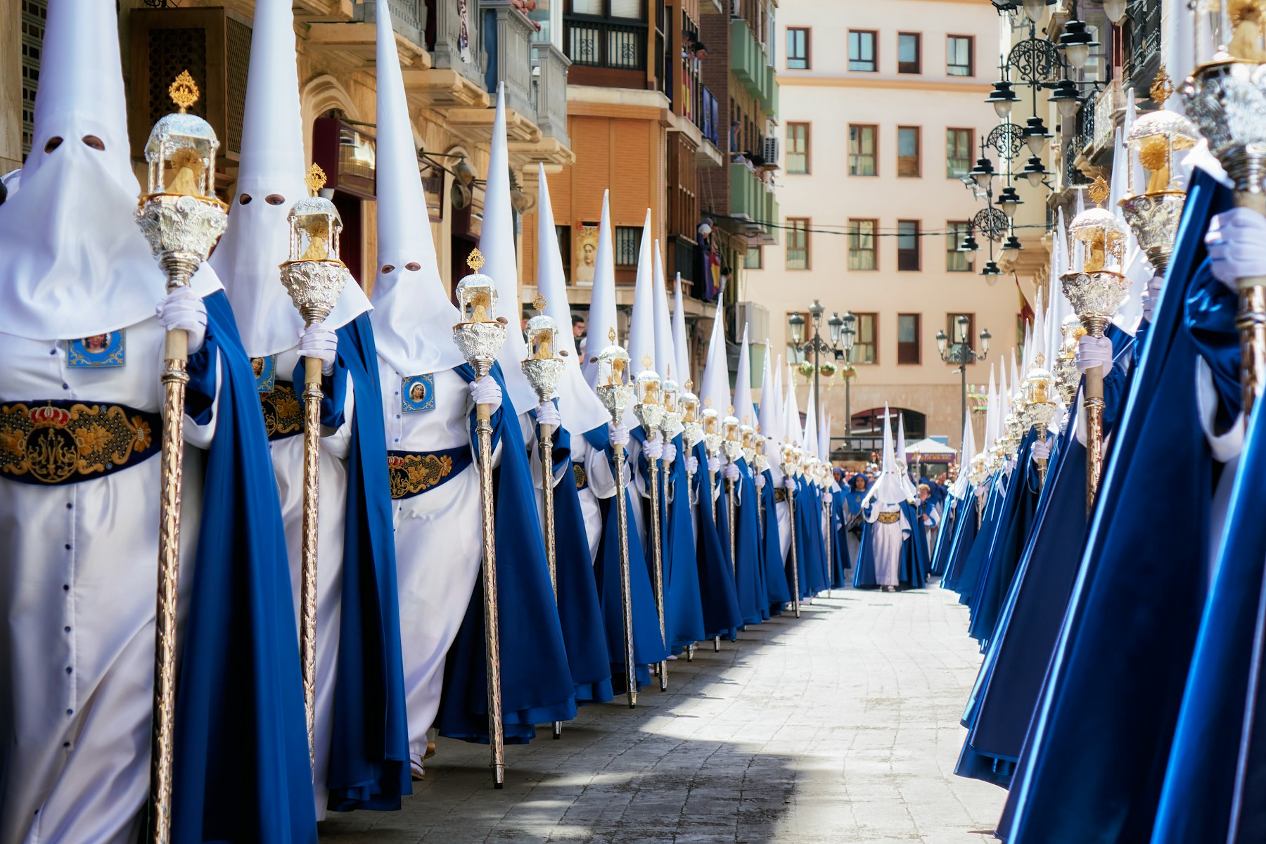 Nazarenos in white capirotes and blue capes carrying ornate staffs in a Semana Santa procession