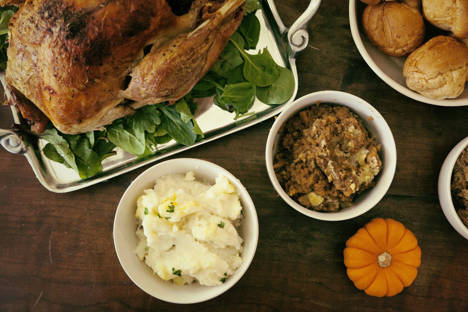 A Thanksgiving table seen from above with roast turkey, stuffing, mashed potatoes, bread rolls and a small pumpkin