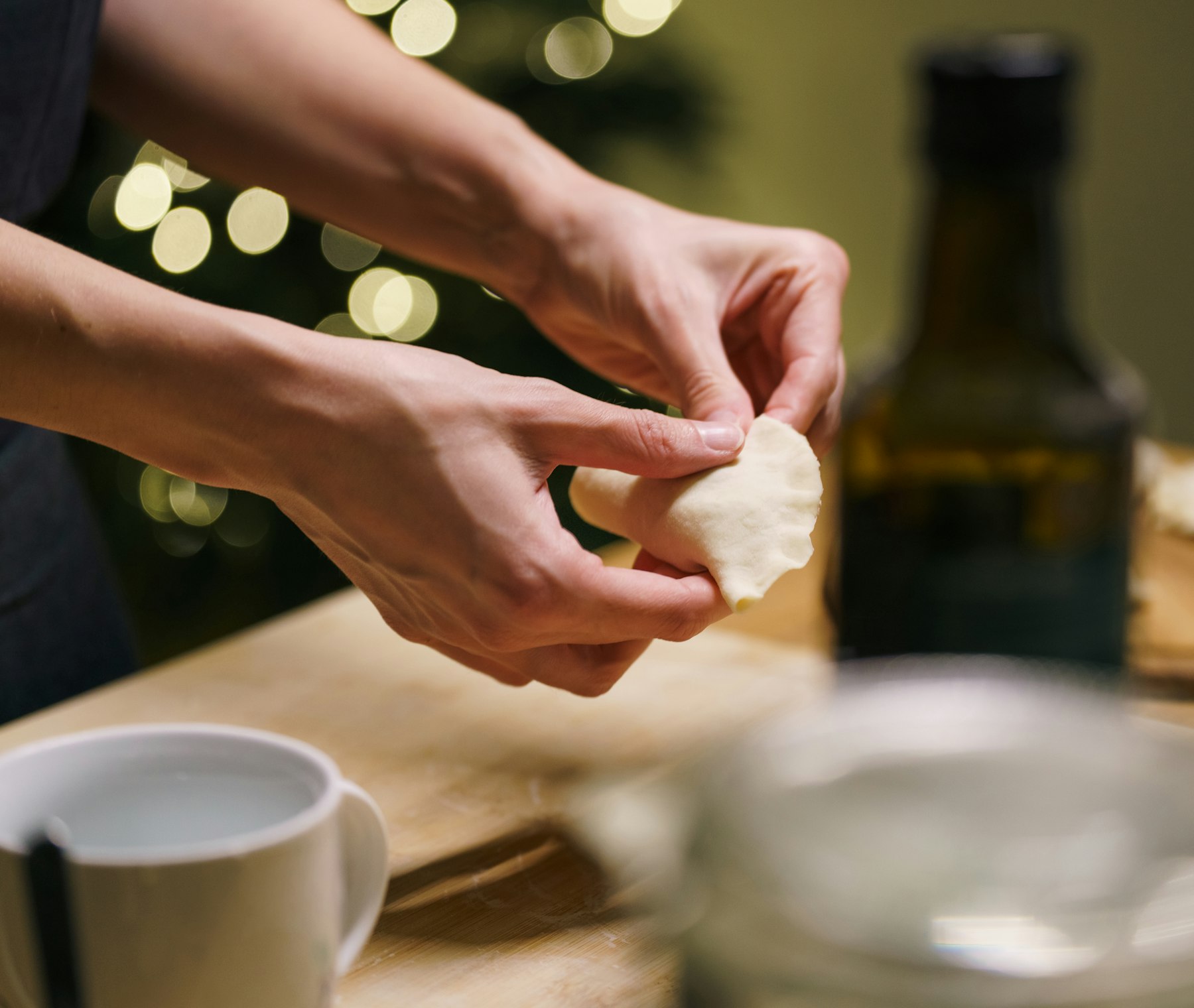 Hände drücken am Küchentisch die Ränder einer selbst gemachten Pirogge zusammen, im Hintergrund leuchten die Lichter eines Weihnachtsbaums
