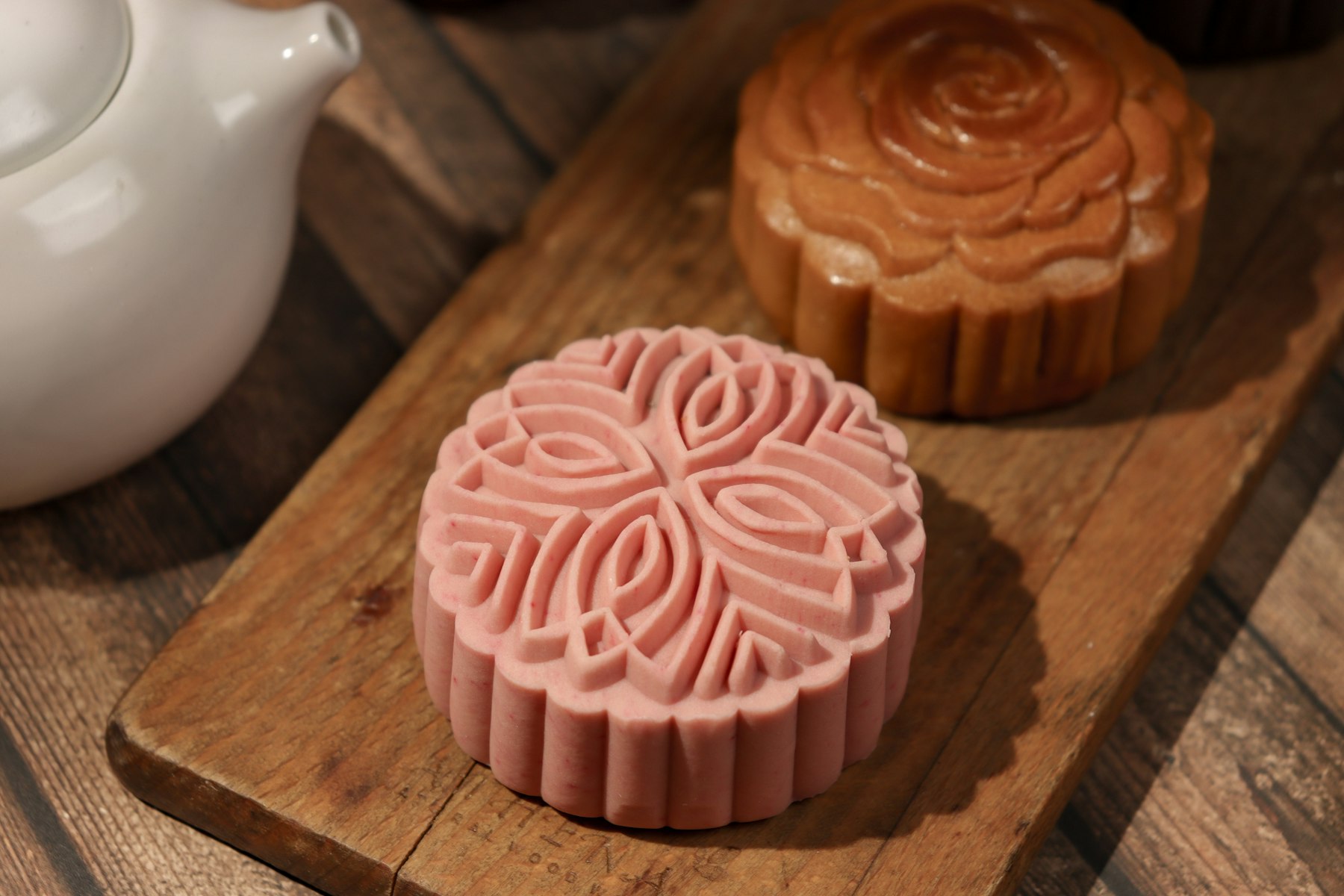 A pink snow-skin mooncake and a baked mooncake on a wooden board beside a teapot