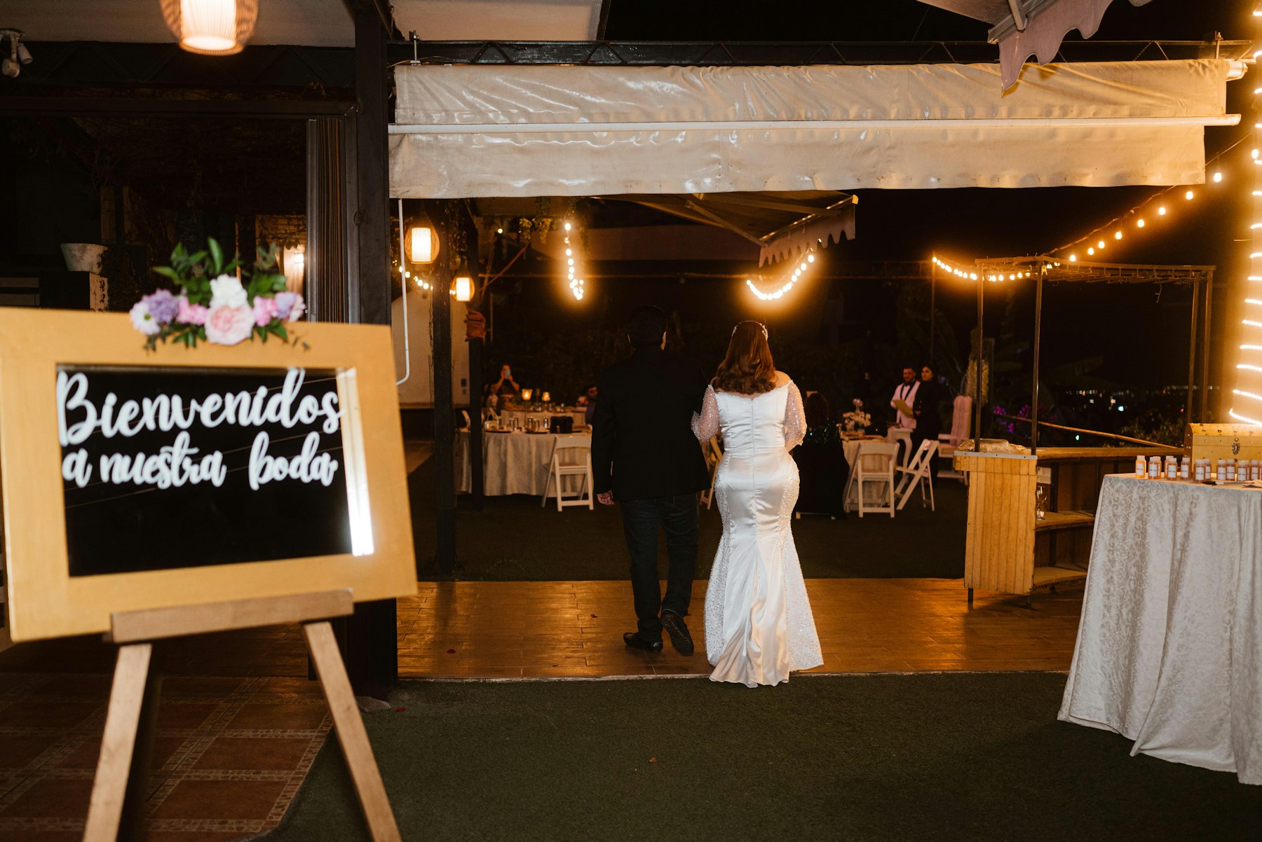 A chalkboard reading “Bienvenidos a nuestra boda” at the entrance to an evening wedding reception as the bride and groom walk in